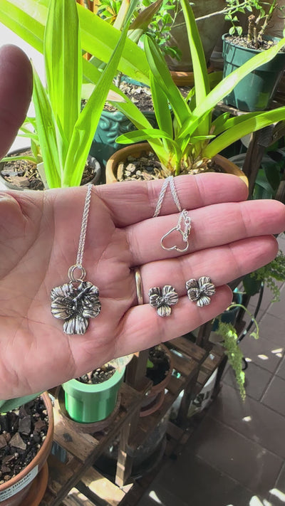 A video showing a jewelry set of sterling silver round-leaved violet earrings and a necklace. There are plants in the background.
