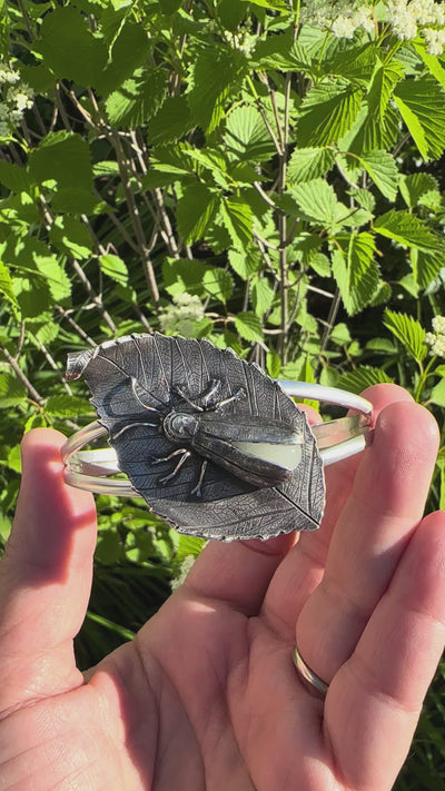 A video showing the glow in the dark sterling silver firefly outdoors. Someone is holding it in front of green bushes. 