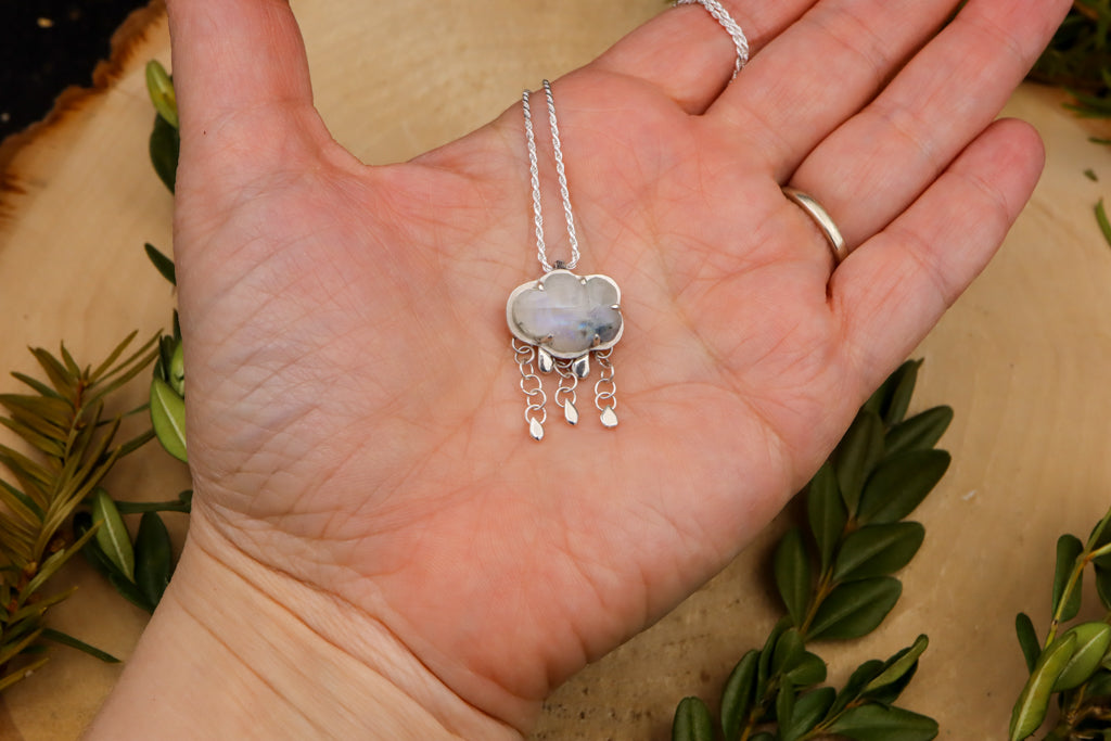 A hand is shown holding the rainbow moonstone necklace to show size. The necklace is about 1 inch across and made entirely from sterling silver. 