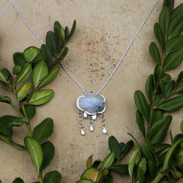 A moonstone raincloud necklace is shown on a necklace on a piece of light tank wood and surrounded by real green branches. 