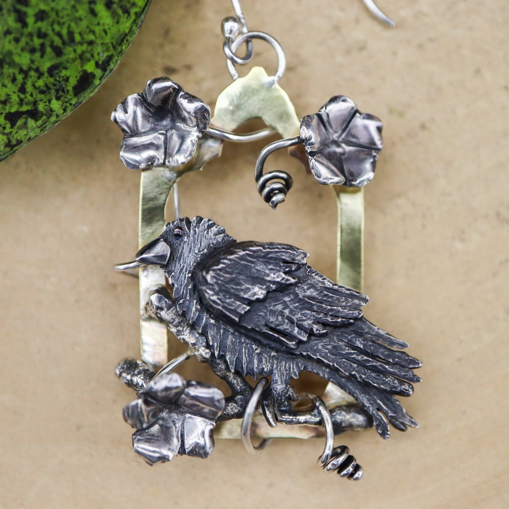 A close up photo of one of the sterling silver raven earrings. You can see the vines of moonflowers and the brass gothic shaped background of the dangle earring.
