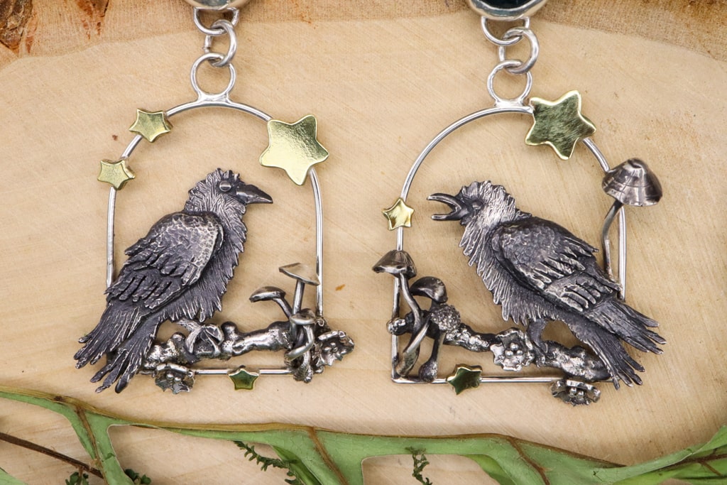A closer view of the handmade sterling silver raven dangle earrings. There are brass gold stars, mushrooms, and crab apple branches and blossoms. They are shown on a piece of light tan wood. 