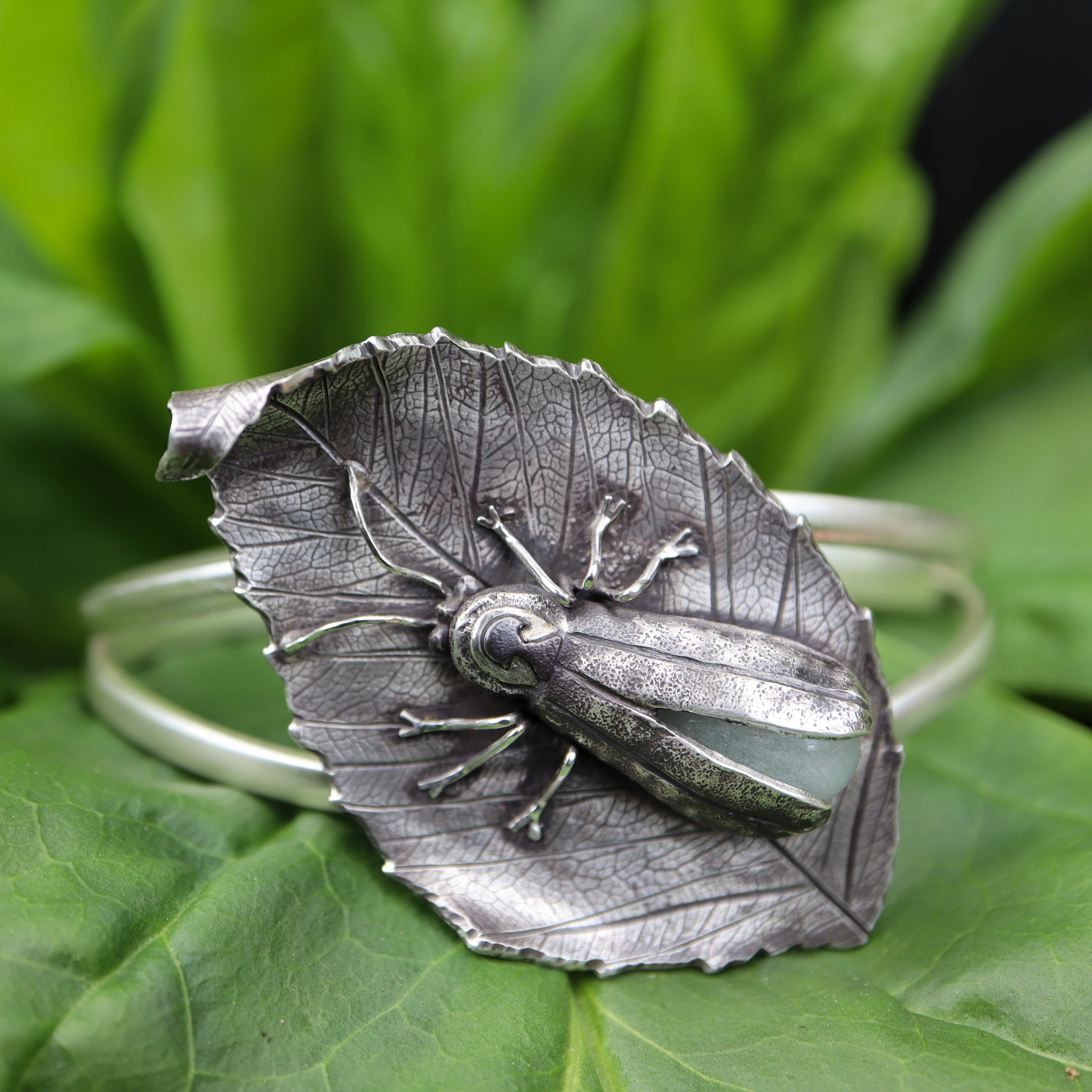 Silver beech leaf-shaped bracelet with a detailed glow in the dark glass abdomen firefly on green leaves.