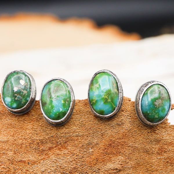 Sonoran Gold Turquoise earrings studs are shown close up with their greens and teals. There are four earrings next to each other on a piece of light brown wood.