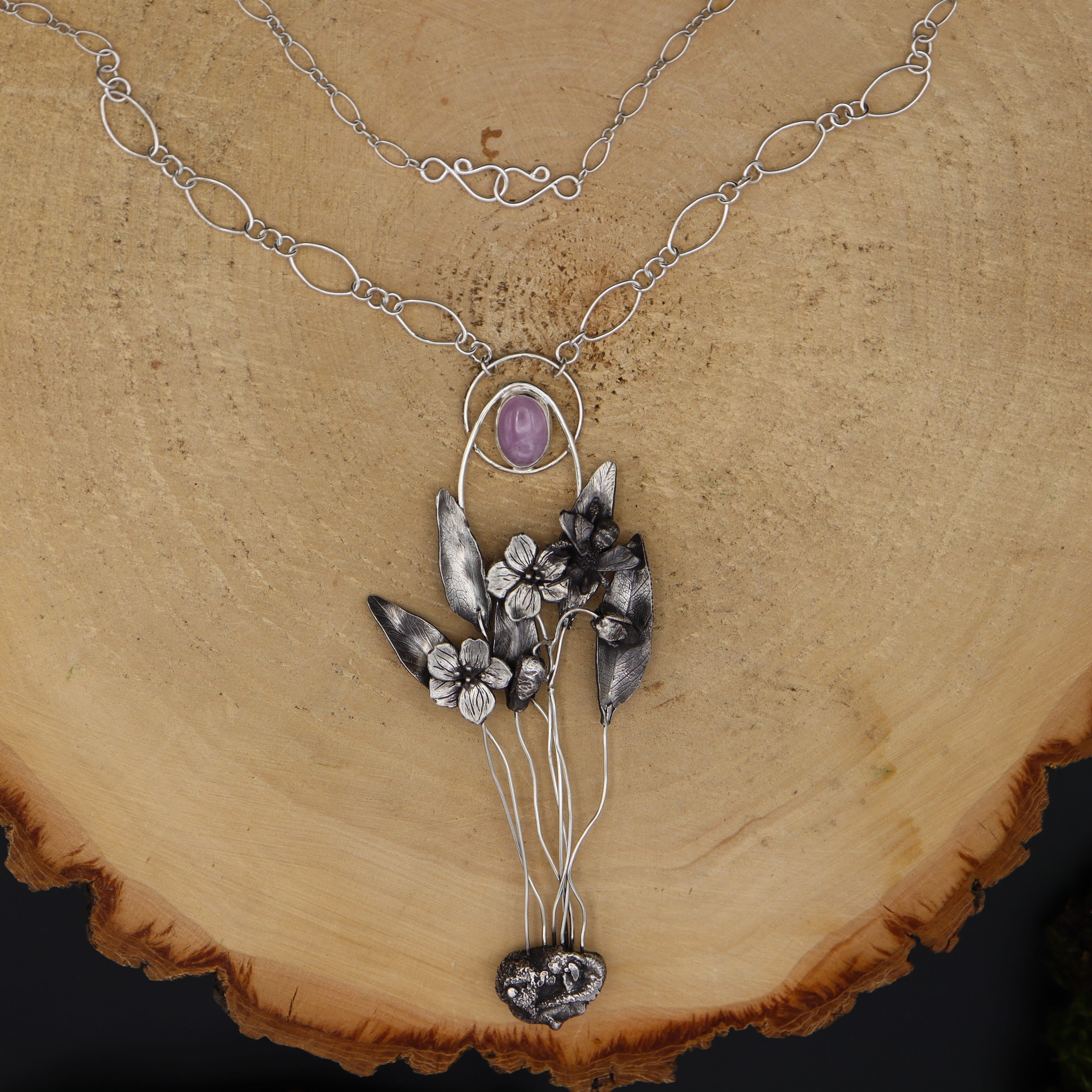 A handmade spring ephemeral wildflower necklace featuring a spring beauty mining bee. There is a light pink kunzite gemstone at the top and it is shown on a light tan piece of wood. 