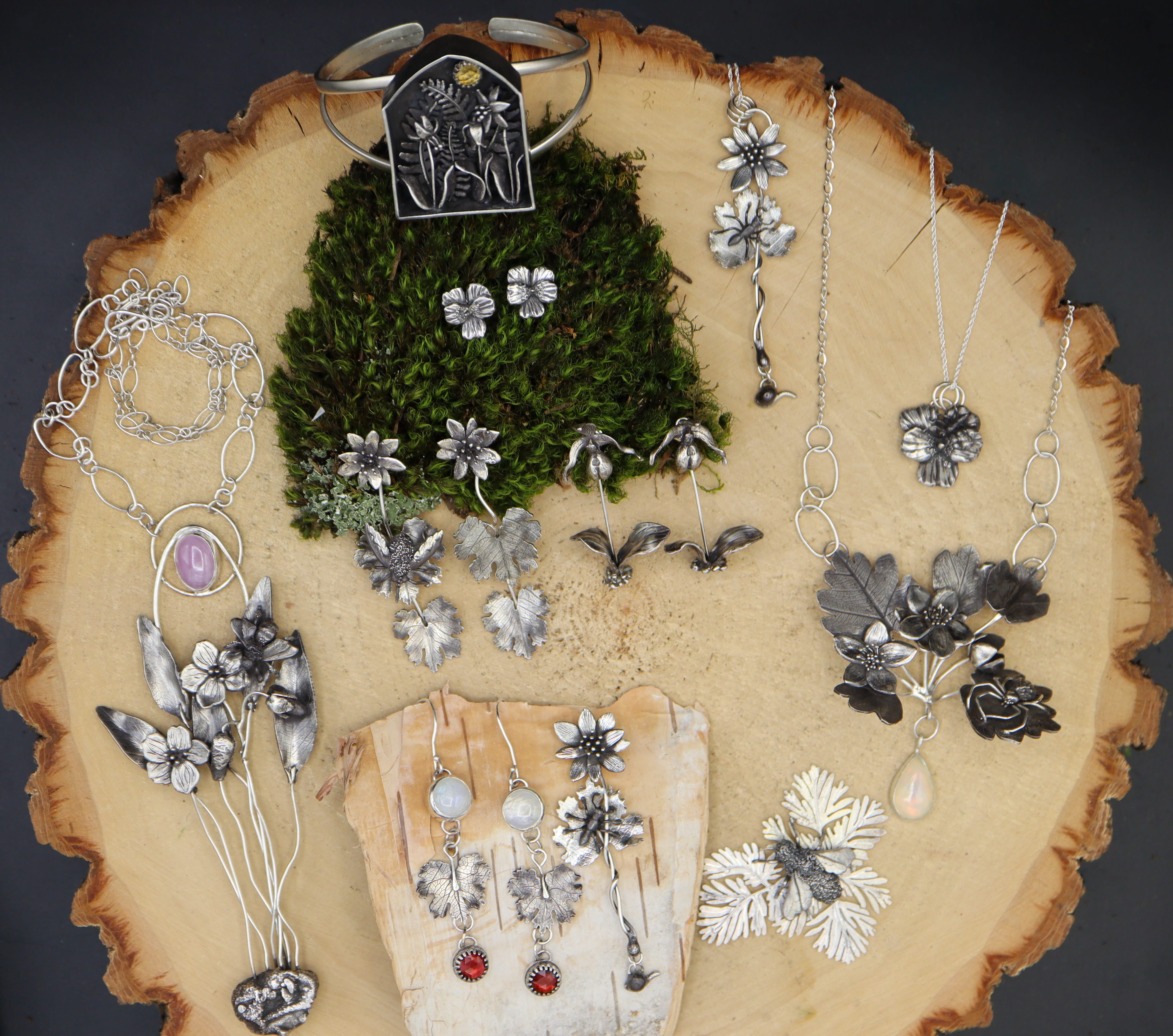 A collection of spring ephemeral wildflower and insect jewelry is shown on a pieces of light tan wood. 
