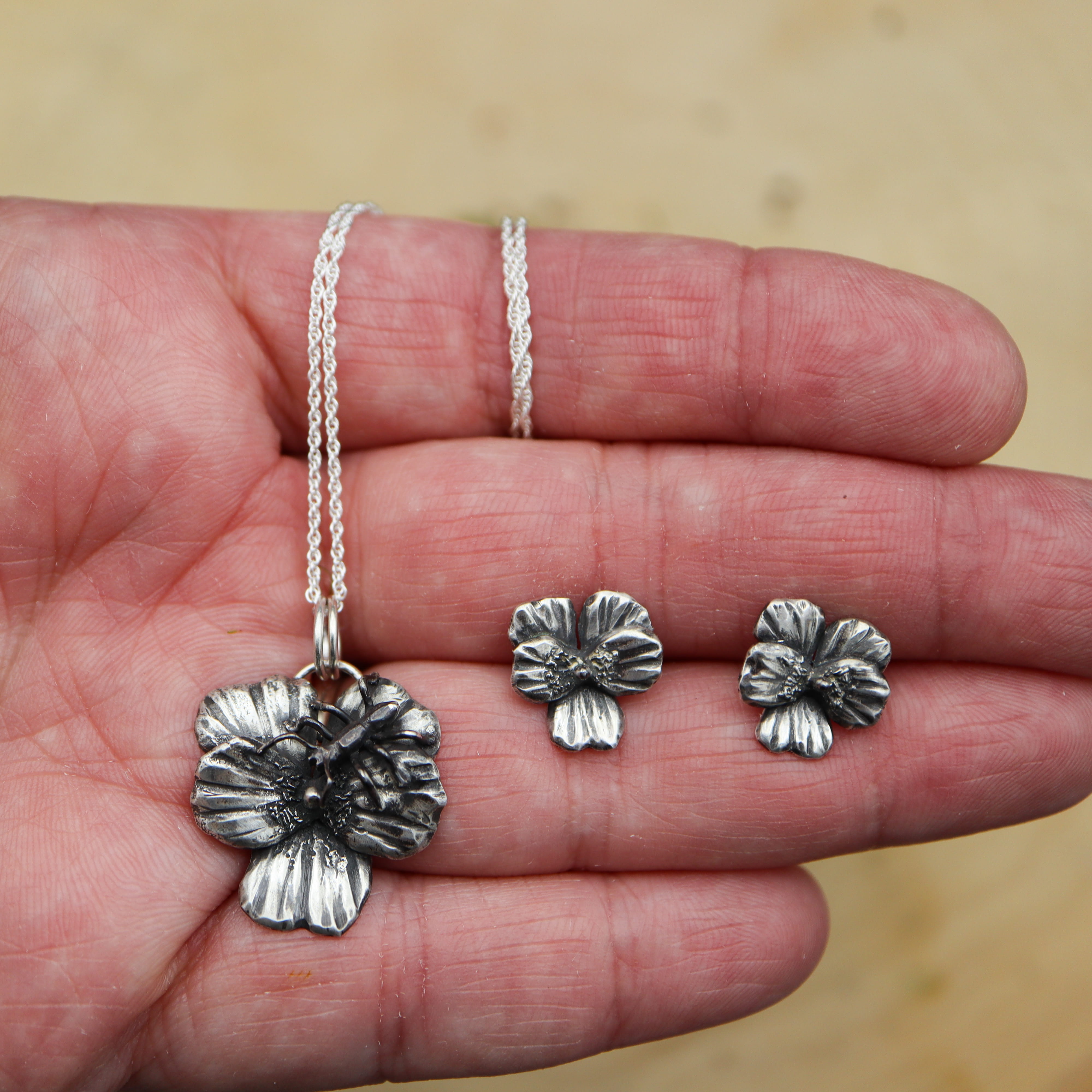 A hand is shown holding the round-leaved violet necklace and earring stud set. 