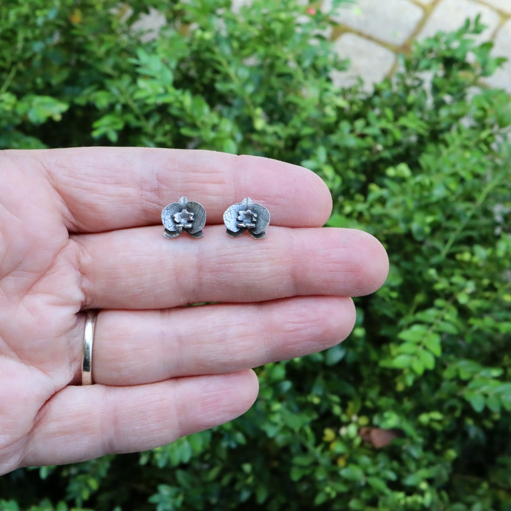 A hand is shown holding the dainty sterling silver orchid flower earrings studs for size reference. They are about 1/2 inch wide. 