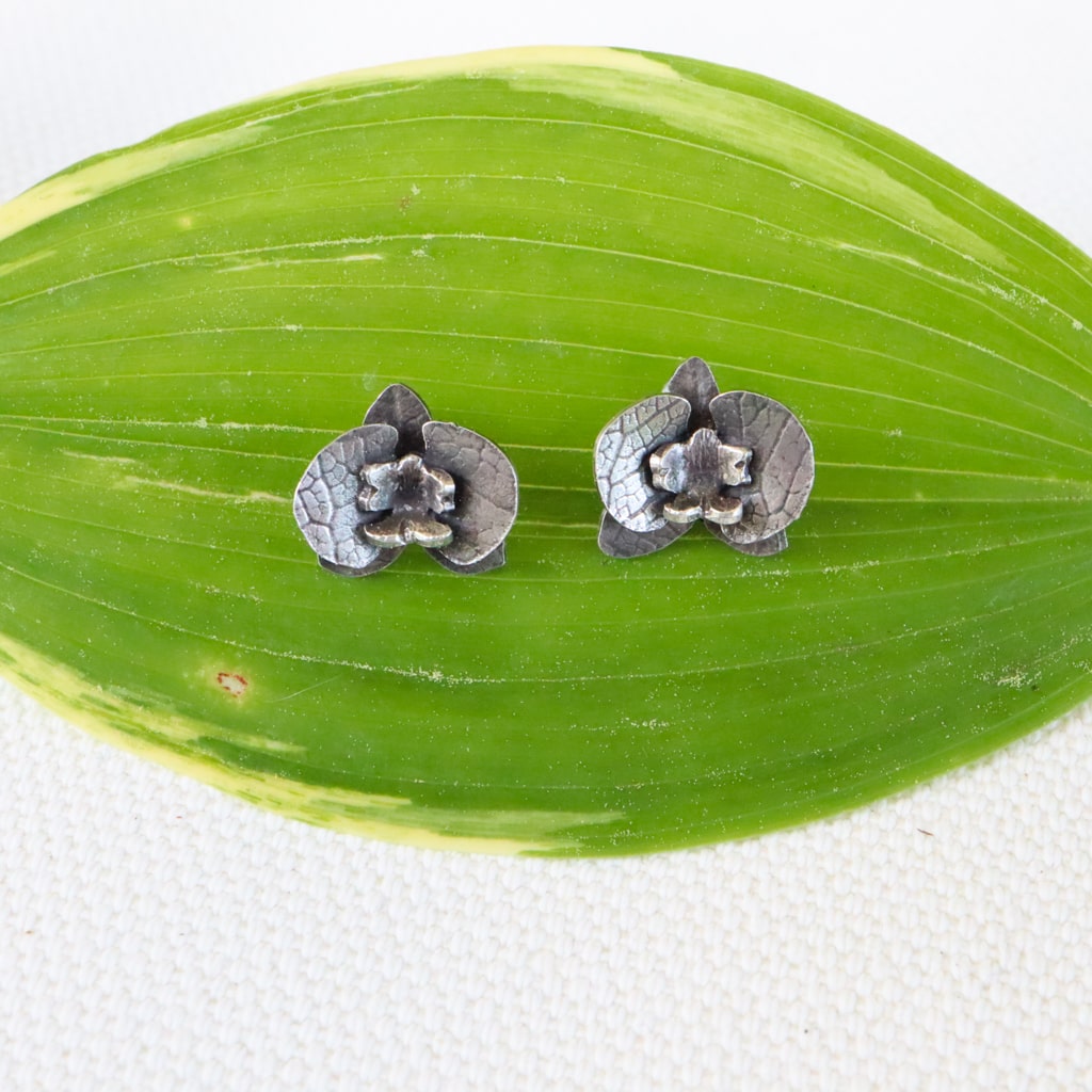 Tiny handmade sterling silver orchid flower earrings are shown on a light green leaf with a white background. 