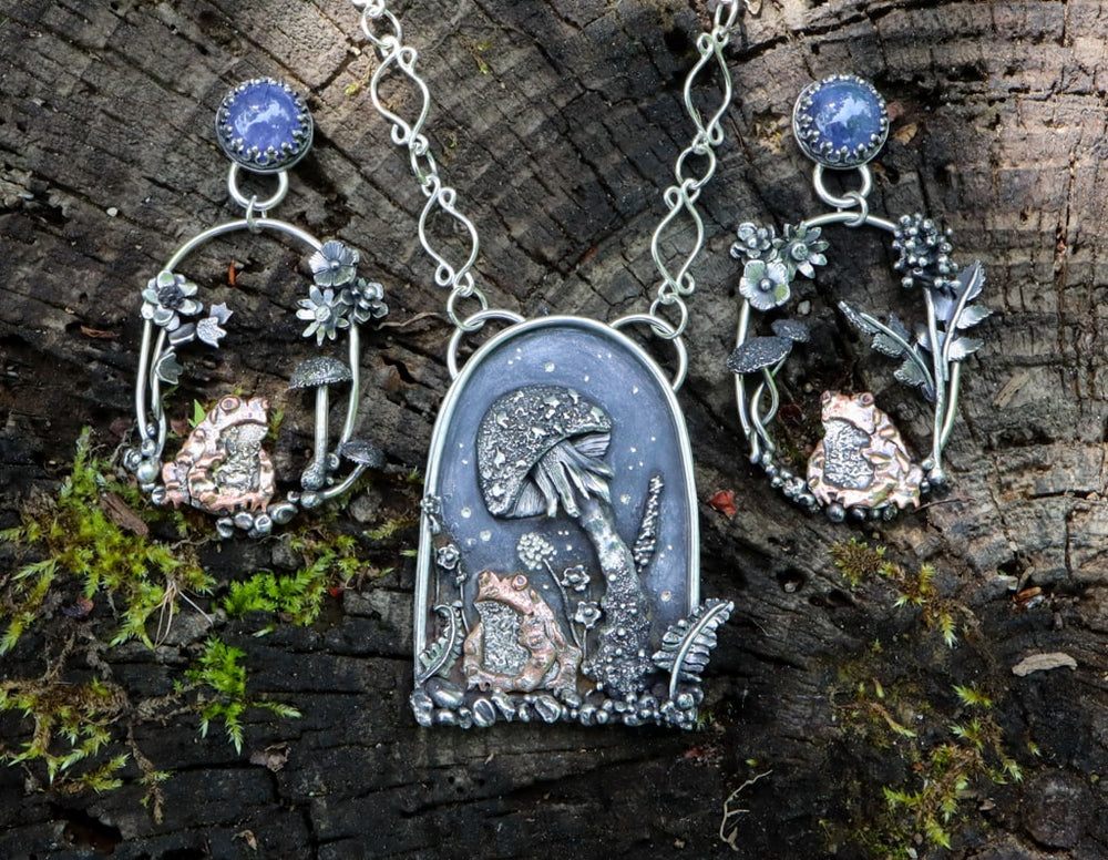 A necklace and pair of earrings all seeming to match with tiny copper toads on each of them. They are all shown in a group on top of a dark piece of wood.