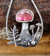 A close up of one of the watermelon tourmaline mushroom earrings showing the bright pink stone at the top of the mushroom. There are ferns, mushrooms, and leaves surrounding the main central mushroom. 