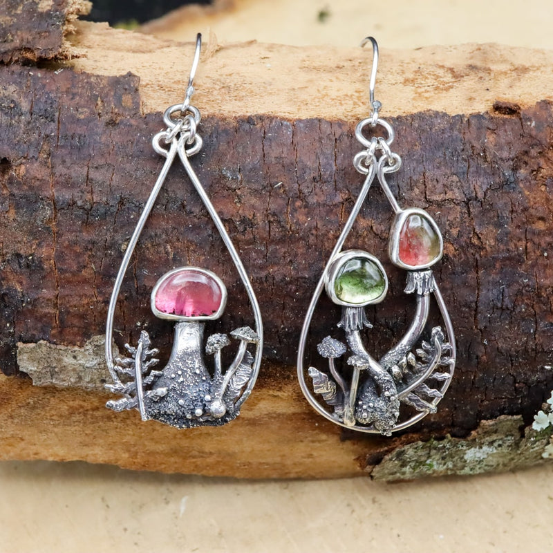 A pair of mismatched watermelon tourmaline mushroom dangle earrings are shown on a piece of darn brown wood. Each earring has a gemstone capped mushroom as the focal point. 