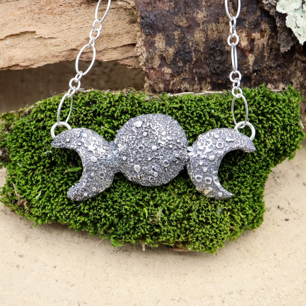 A triple goddess moon necklace is shown on a dark green piece of moss. 