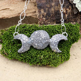 A triple goddess moon necklace is shown on a dark green piece of moss. 