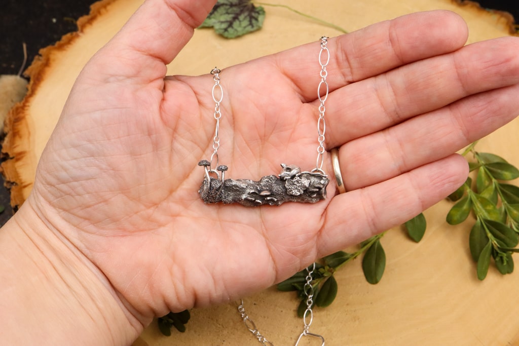 A hand is shown holding the sterling silver mouse and mushroom necklace. 