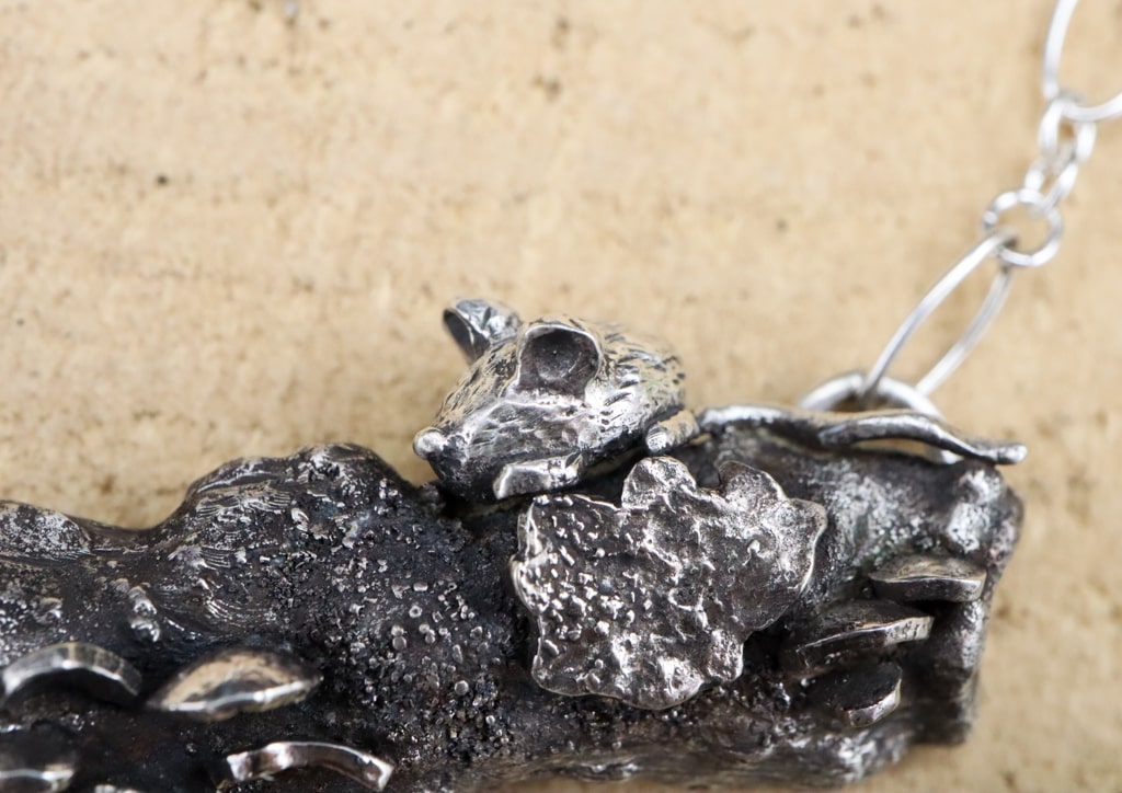 A close up photo of the hand carved sterling silver mouse sitting above a tiny piece of silver lichen. 
