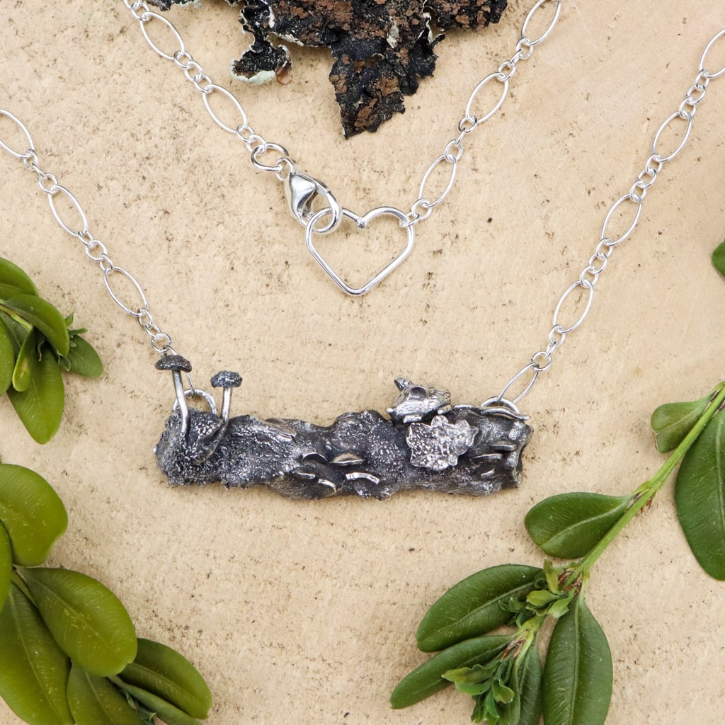 A handmade sterling silver necklace that resemble a forest log. On the silver log there is a tiny silver mouse sitting above some lichen, turkey tail mushrooms, and some button mushrooms. The necklace is shown on a piece of light tan wood and surrounded by greenery. 