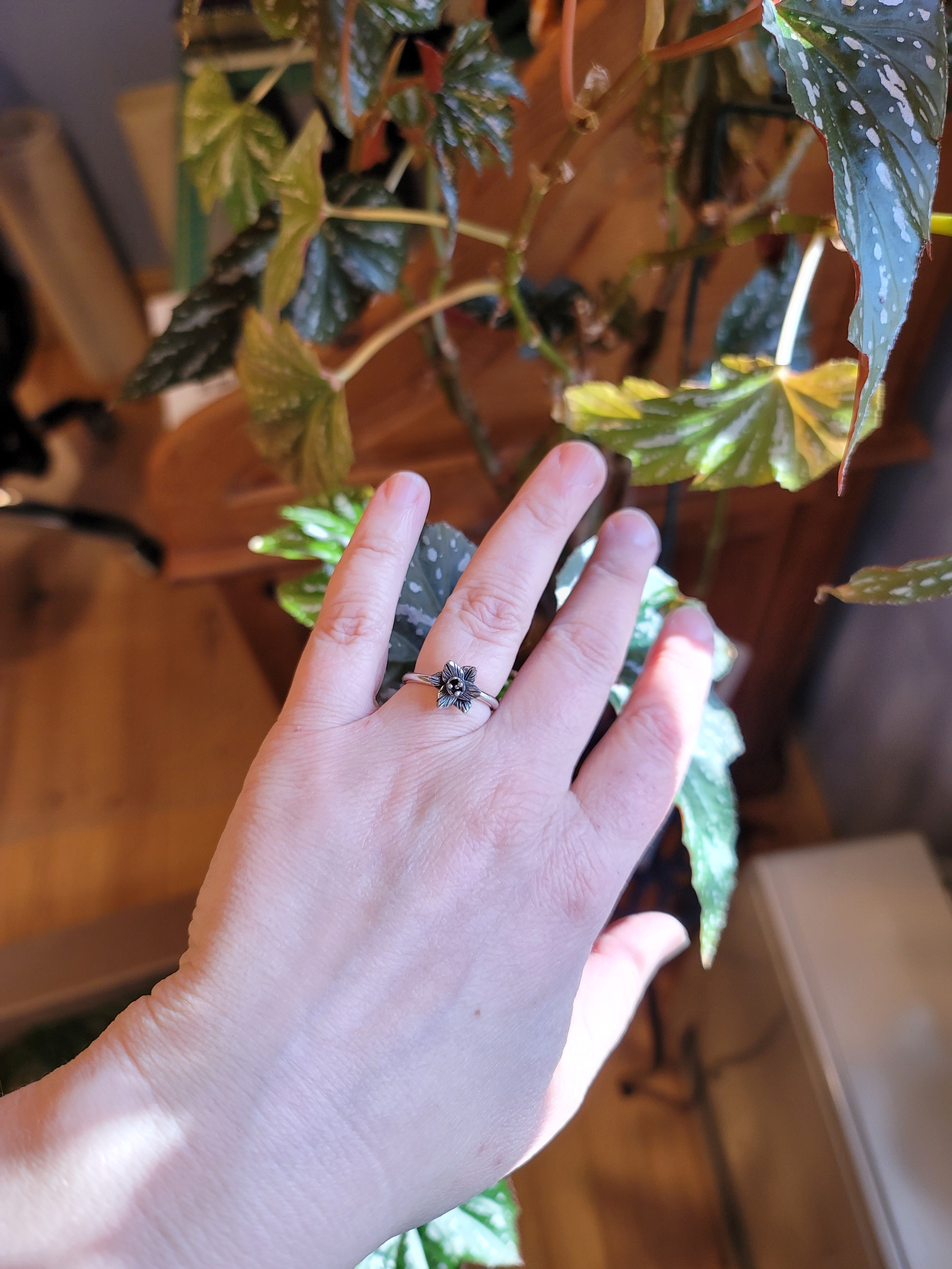A hand is shown wearing the dainty silver daffodil ring. It is in front of a dark green begonia plant with some sunbeams coming in the back.