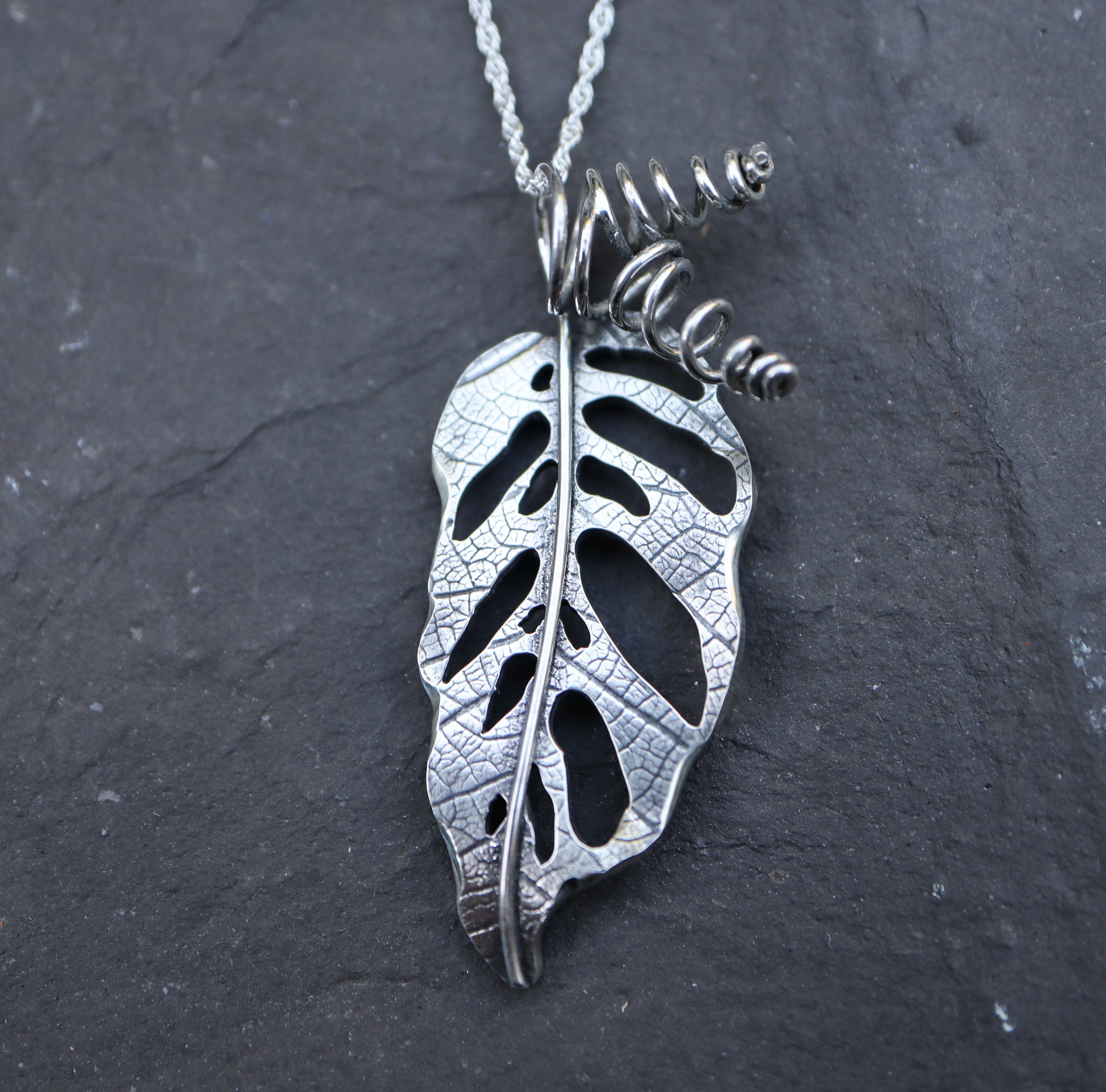 A handmade sterling silver monstera adansonii necklace made by The Striped Cat Metalworks. The necklace is pictured on a dark grey piece of slate. 