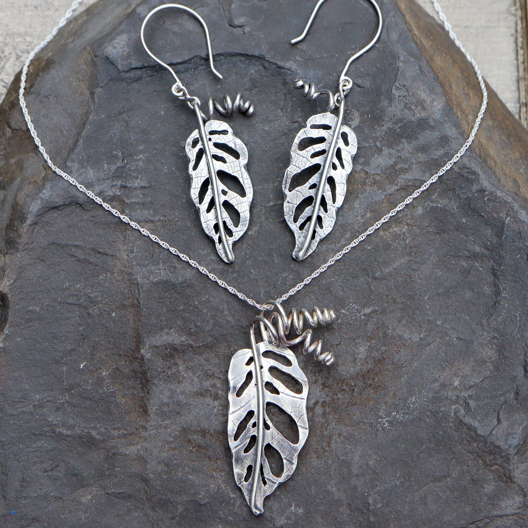 Monstera adansonii earrings and a necklace set that come together. The necklace leaf is about 1.5 inches tall and the earrings are a little smaller. They are shown on a dark grey piece of slate. 