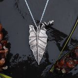 A handmade sterling silver alocasia houseplant leaf necklace. The leaf is about 1.25 inches tall and has a twisty stem holding it onto a necklace. 