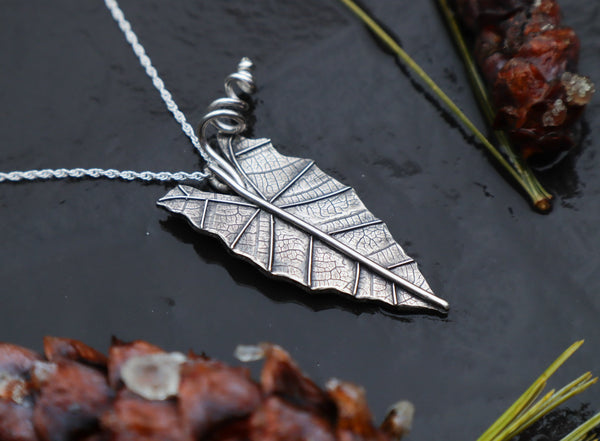 A handmade sterling silver alocasia african mask leaf made into a necklace shown from the side view. It is about 1.5 inches tall and is pictured on a dark slate stone with pinecones and greenery around it.