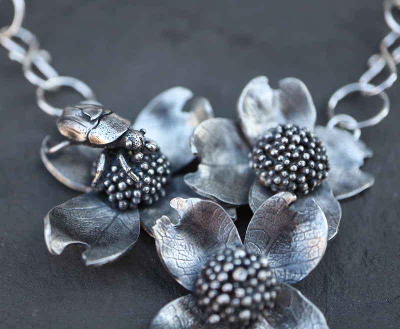 A close up side view of the hand carved sterling silver beetle sitting on top of a dogwood flower. 