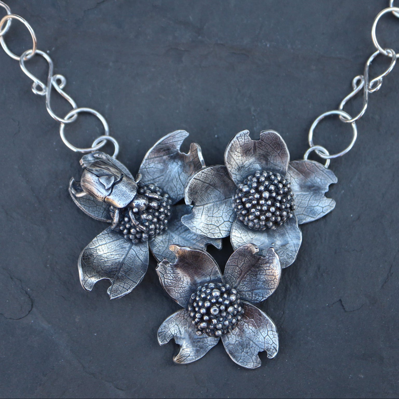 A handmade sterling silver dogwood flower and beetle necklace shown on top of a dark grey piece of slate. 