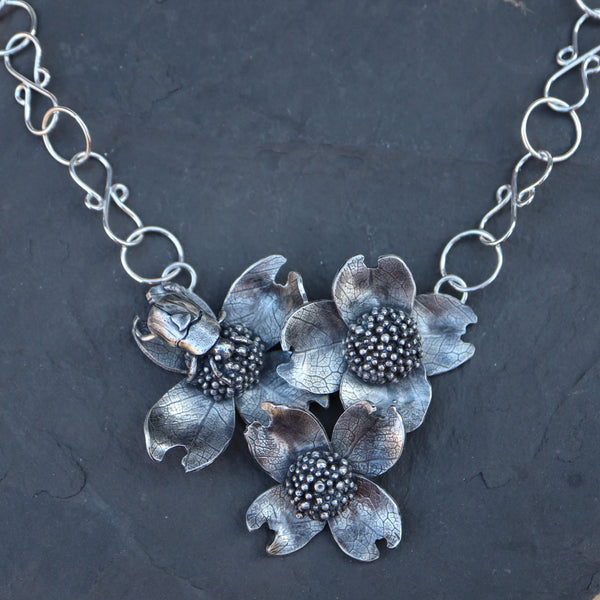 A handmade sterling silver dogwood flower and beetle necklace shown on top of a dark grey piece of slate. 