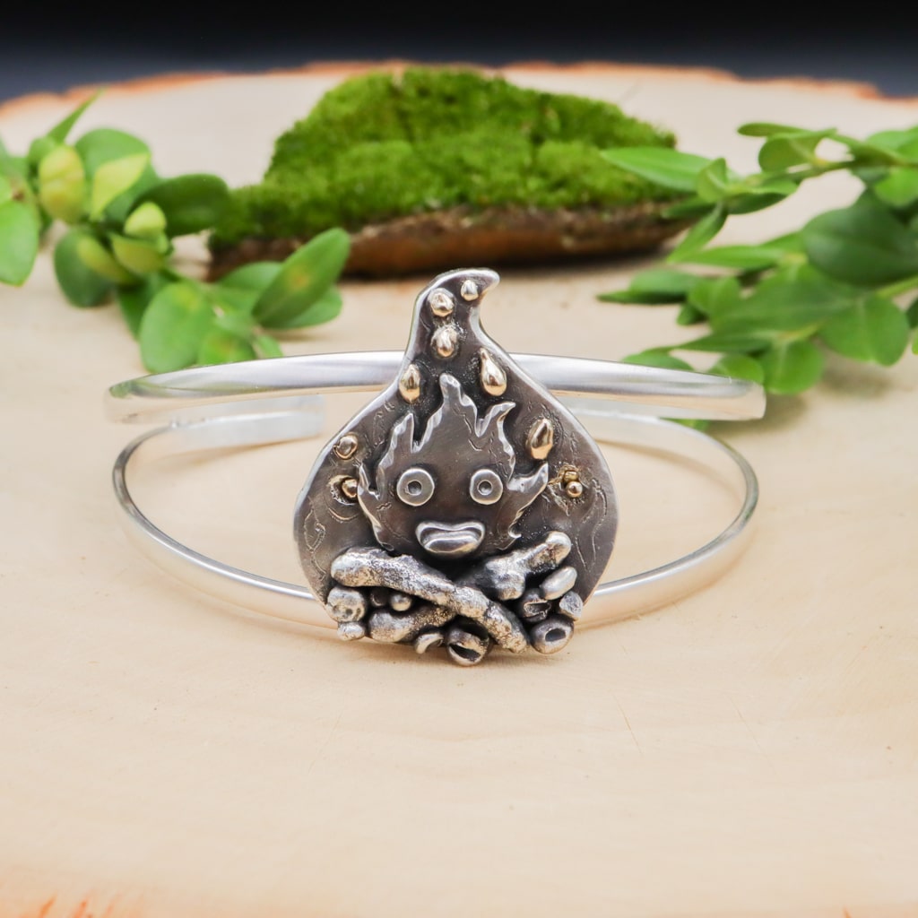 A sterling silver one of a kind Calcifer bracelet. He is sitting in the middle of it with a big smile and flames above him. Some of flames are made with bits of real gold. The bracelet is shown on a light tan piece of wood.