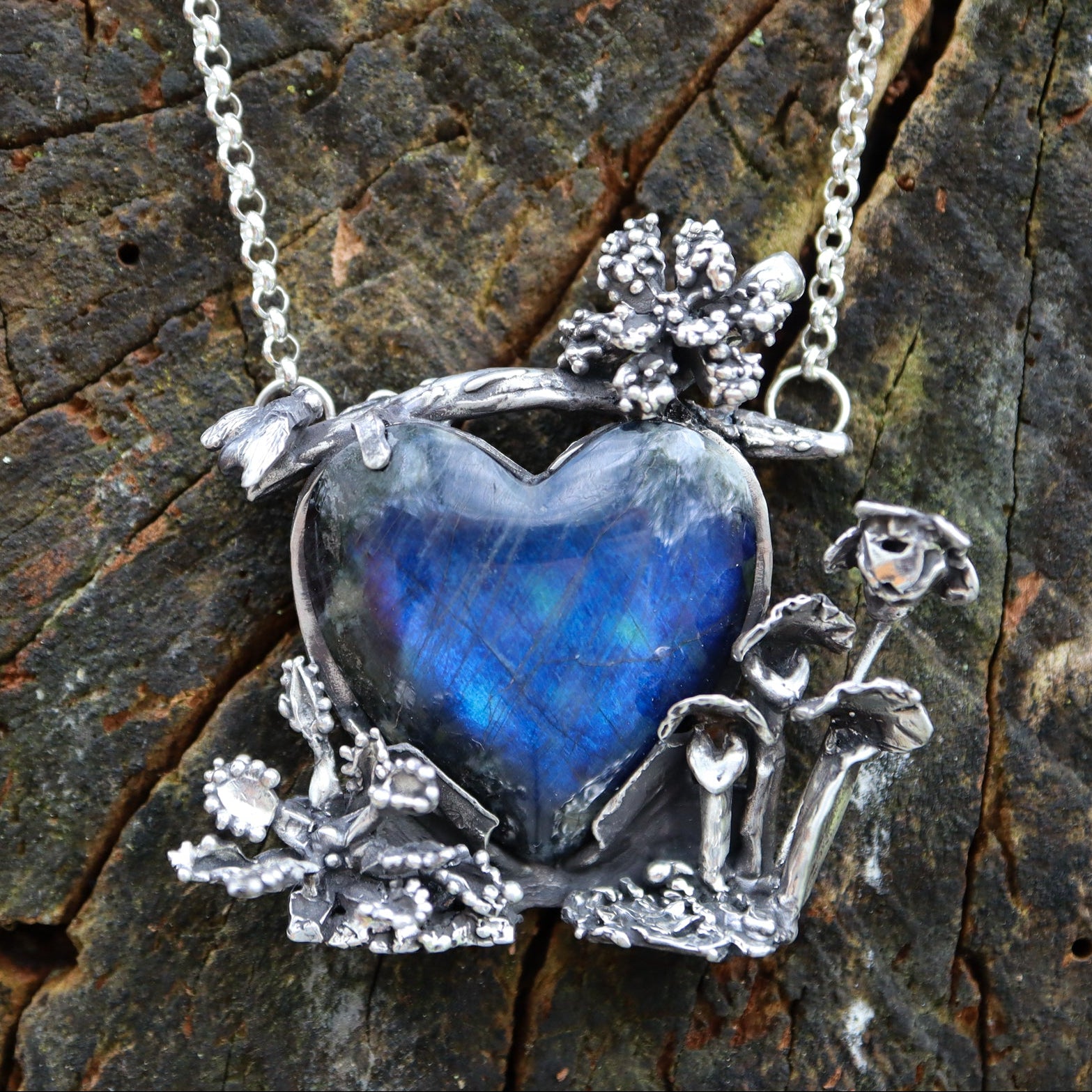 A deep blue spectrolite stone is surrounded by carved silver carnivorous plants with a tiny silver fly on the top left of the pendant. The necklace is shown on a dark brown piece of tree bark. 
