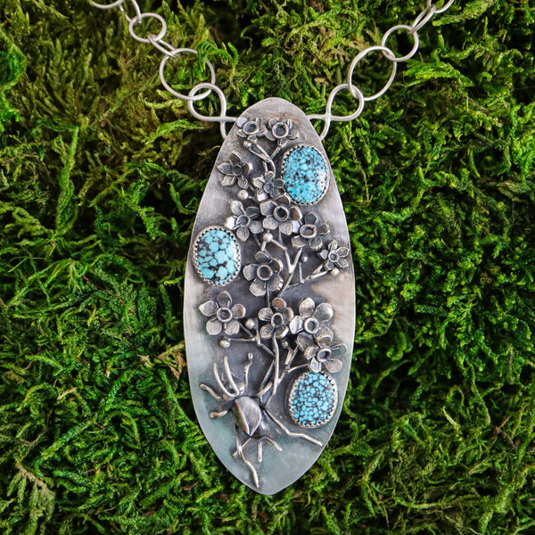 A sterling silver handmade necklace in an elongated shield shape. There is a hand carved cricket at the bottom of the pendant and above her is a sprig of silver forget-me-not flowers. There are three light blue spotted turquoise stones mixed among the flowers. The necklace is shown in a bed of bright green moss. 