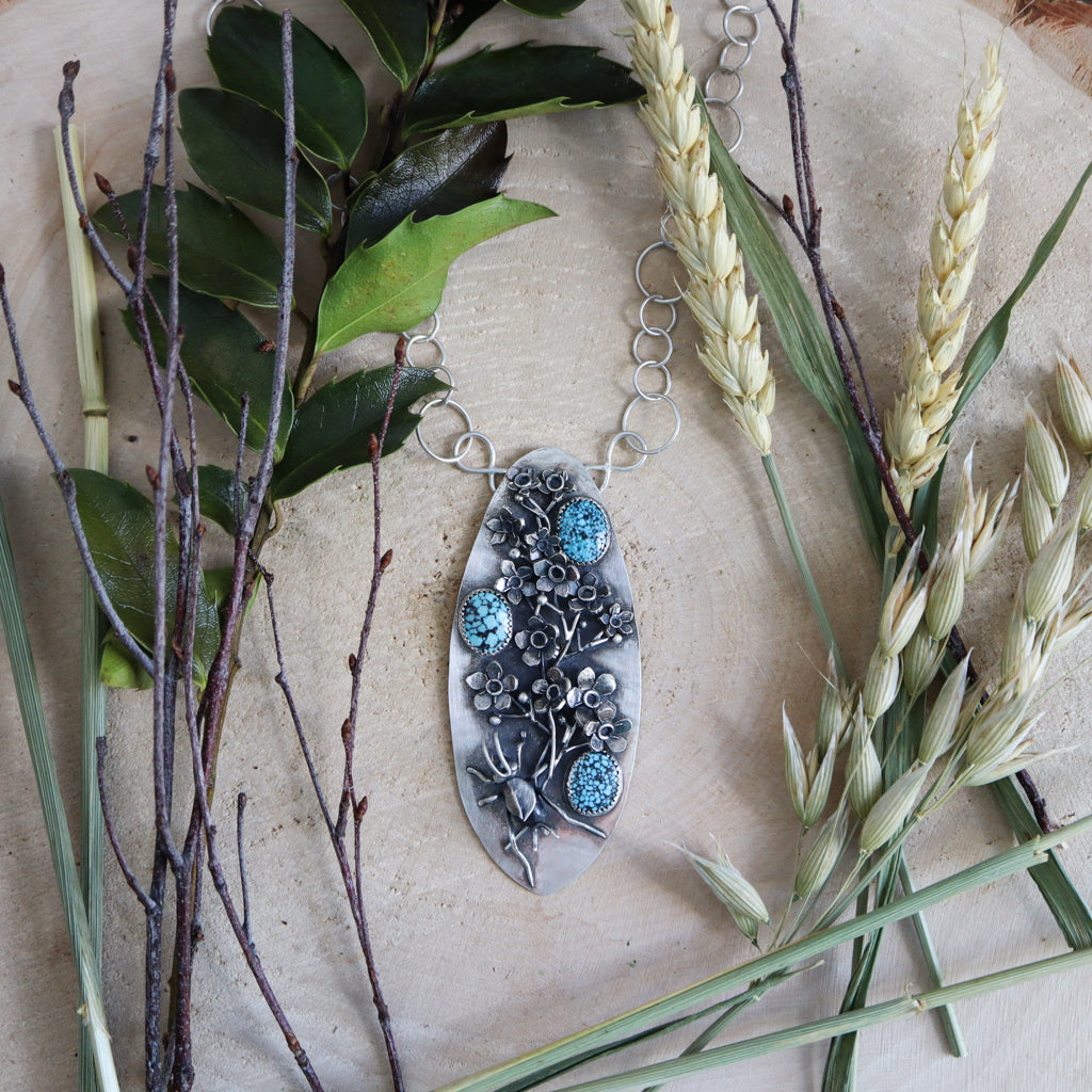 A photo farther away showing the cricket necklace and part of the hand made loop chain. There are sprigs of wheat and holly twigs around the necklace. 