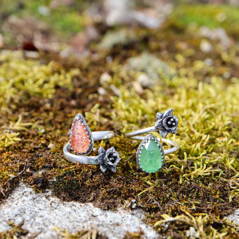 Two handmade sterling silver daffodil rings are shown on some real moss. The right on the left is made with an orange sunstone and the ring on the right is make with a grass green tsavorite garnet. 
