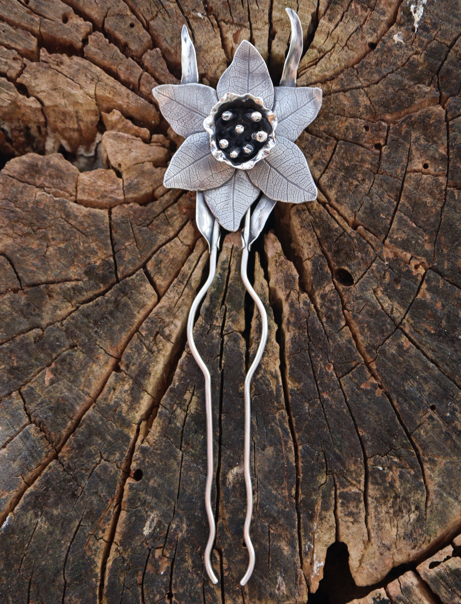 A handmade sterling silver daffodil flower hair pick. It is about 6 inches tall and shown in front of a piece of light brown tree bark. 