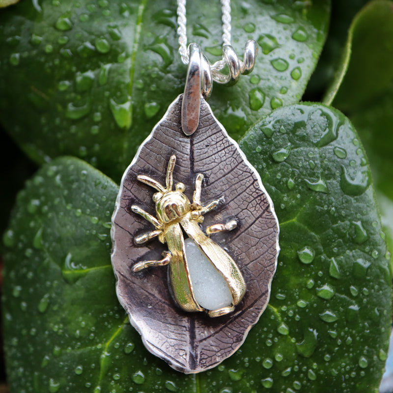 A 14k gold firefly insect is sitting in the middle of a sterling silver leaf. Her abdomen is made from low in the dark borosilicate glass. The necklace is shown on a grouping of dark green leaves with rain drops on them. 