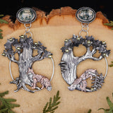 Mismatched sterling silver fox hoop earrings are shown on a light tan piece of wood. The earrings are about 3 inches tall and feature two moss agates at the top of the design. Each of the fox earrings is showing a different forest scene and both are below apple trees. 