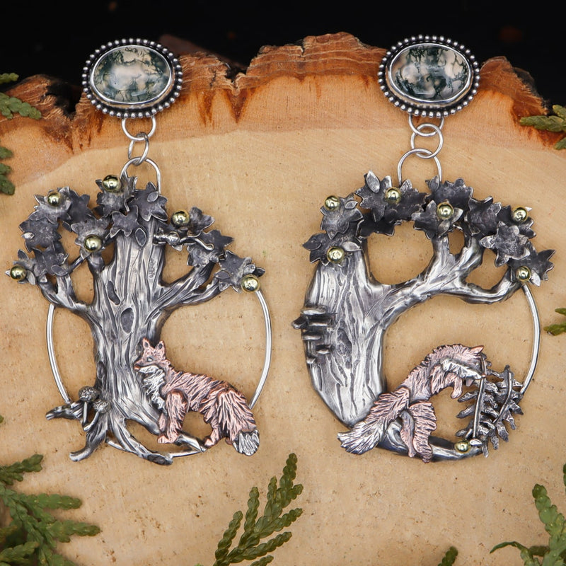Mismatched sterling silver fox hoop earrings are shown on a light tan piece of wood. The earrings are about 3 inches tall and feature two moss agates at the top of the design. Each of the fox earrings is showing a different forest scene and both are below apple trees. 