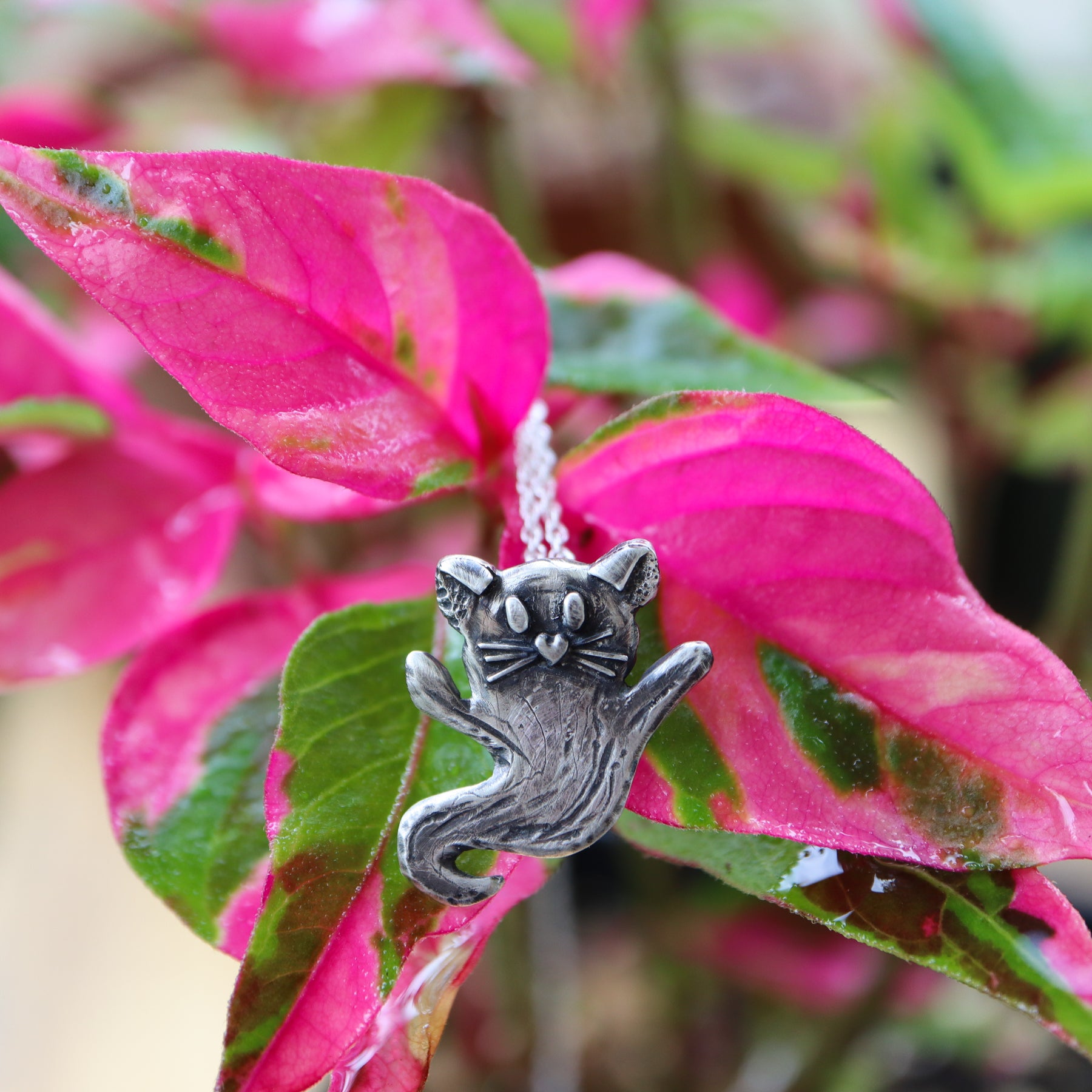 A sterling silver handmade necklace in the shape of a ghost but made to look like a cat. She has little ears on top, two evey, a heart shaped nose, and whiskers. The necklace is shown in front of a bight hot pink real plant.