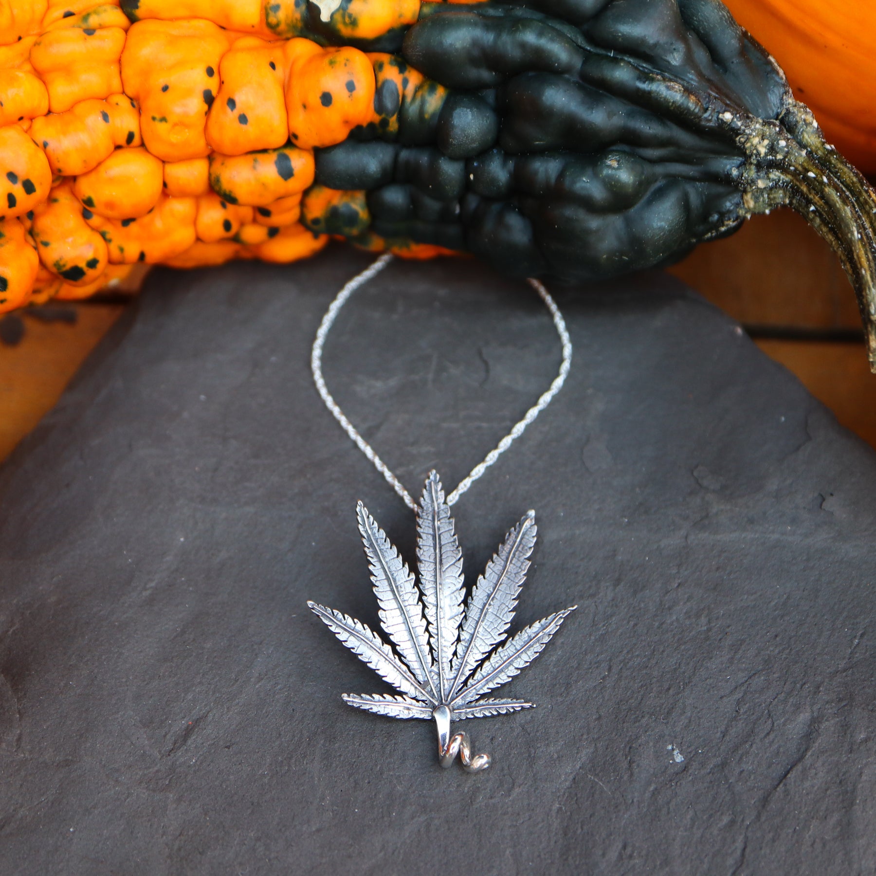 A sterling silver handmade cannabis leaf pendant necklace is about 1.25 inches tall and shown on a dark grey piece of slate. There are orange and green gourds around the necklace.
