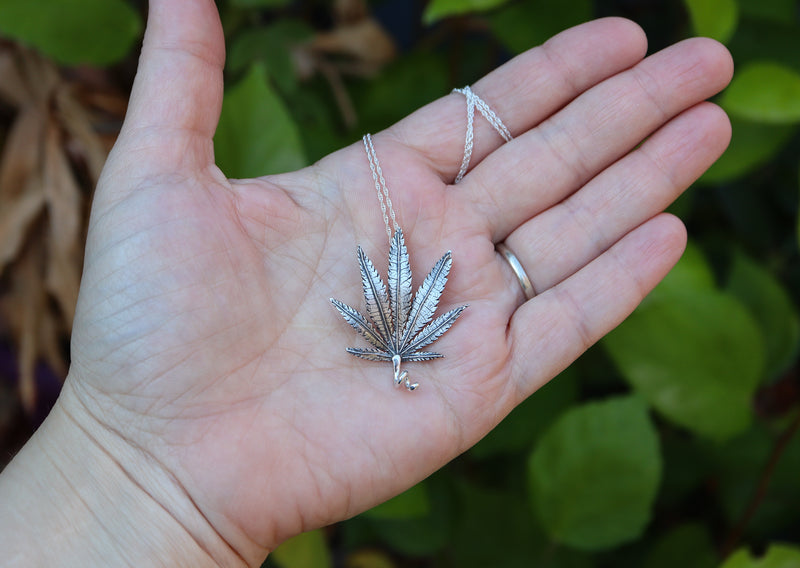 A hand is shown holding a handmade sterling silver cannabis leaf necklace to show the size. It is on an 18 inch long necklace. 