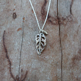 A dainty sterling silver monstera adansonii necklace charm is shown on a piece of light brown and dark brown wood.