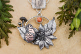 A close up photo of the monarch butterfly necklace shown with real greenery next to it and on a light tan piece of wood.