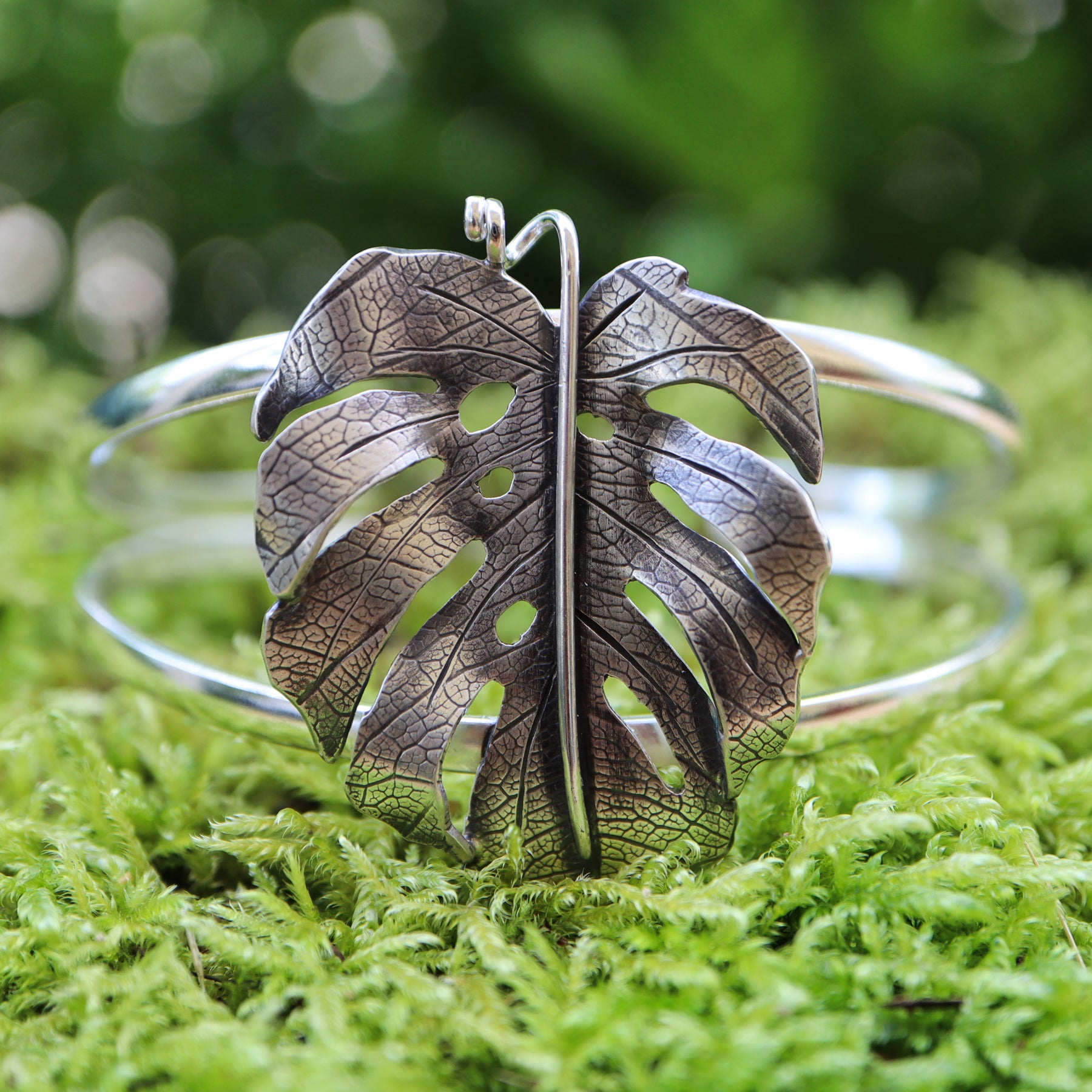 A sterling silver monstera deliciosa leaf bracelet is shown on a piece of light green moss. The leaf is about 1.5 inches tall and has a little curly stem at the top. 