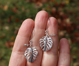 A hand holding a pair of monstera deliciosa dangle earrings with green bushes in the background.