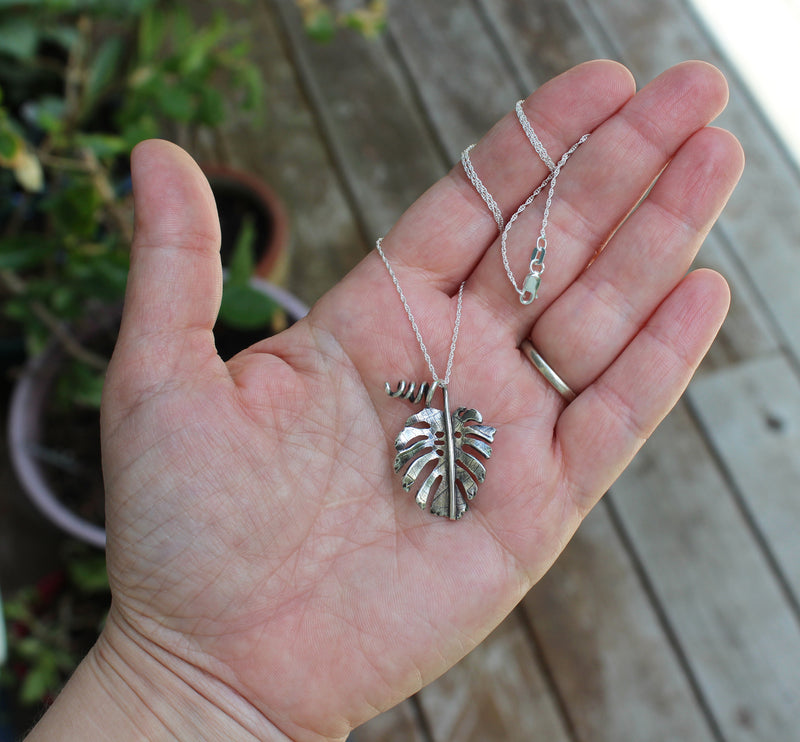 A hand is holding the monstera deliciosa sterling silver necklace to show size. 