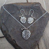 Monstera deliciosa necklace and earring set made from sterling silver from The Striped Cat Metalworks. The jewelry is shown on top of a dark grey slate.