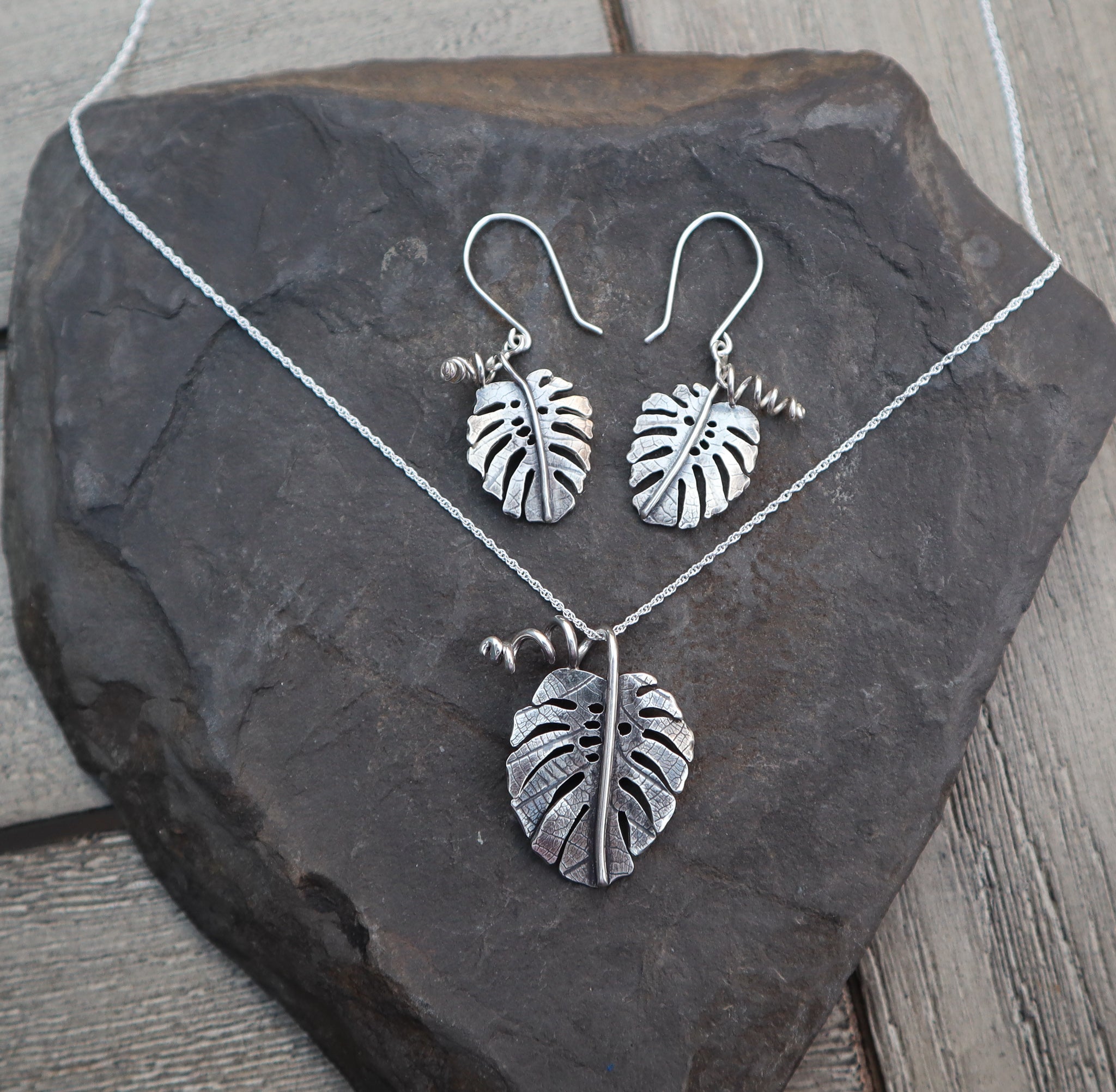 A monstera deliciosa earring and necklace set shown on a piece of dark grey slate stone. 