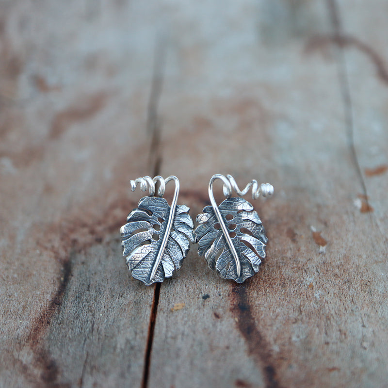 Handmade sterling silver monstera deliciosa stud earrings. They are about 1/2 inch tall and have twisty little stems at the top of the leaf. They are shown on a piece of light brown wood.