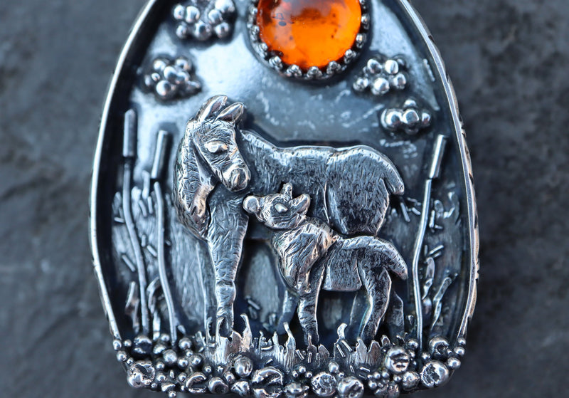 Sterling silver handmade mother moose and baby pendant made by The Striped Cat Metalworks. The pendant has an amber colored stone and shown on a slate piece of rock. 